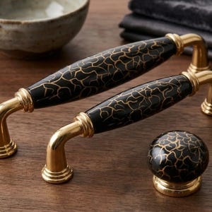 May include: Two elongated cabinet pulls and one round cabinet knob featuring a glossy black ceramic center with a gold crackle pattern. Each piece is finished with polished gold metal hardware at the mounting points and base.
