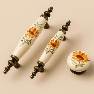 Puede incluir: Juego de herrajes para armario de color crema con dos tiradores alargados y un pomo redondo. Cada pieza está adornada con un diseño de girasol pintado a mano en tonos naranja, amarillo y verde, complementado con detalles metálicos en tono bronce antiguo.