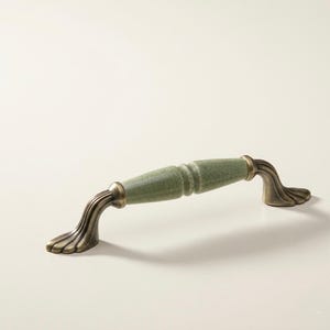 Puede incluir: Un tirador de armario con un centro de cerámica verde claro craquelado y herrajes de metal color latón antiguo. El tirador tiene un diseño curvo con detalles decorativos, adecuado para cajones o armarios.