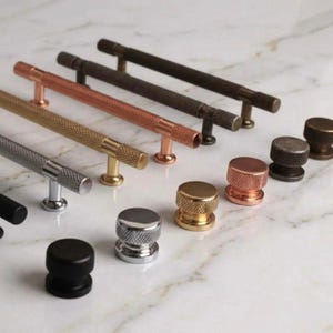 May include: A collection of cabinet hardware in various metallic finishes, including silver, gold, copper, and black. The selection features both bar pulls and knob styles, all with textured surfaces. The hardware is arranged on a white marble surface.