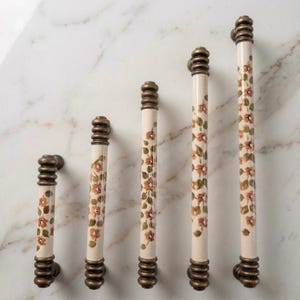 May include: Five cream-coloured cabinet pulls with a floral design in orange and green. The pulls have bronze-coloured bases and tops with a ribbed design. The pulls are arranged in a row, varying in length. The longest pull is approximately 20 cm.