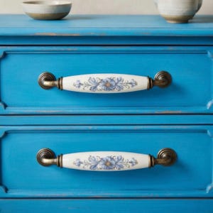 Puede incluir: Primer plano de una cómoda azul brillante con dos cajones. Cada cajón tiene un tirador de cerámica blanca con un diseño floral en azul y dorado. Los tiradores están sujetos con herrajes de latón antiguo. Dos cuencos de cerámica encima.