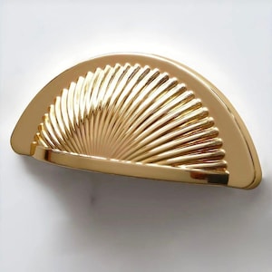 May include: A gold-colored, half-moon shaped cabinet pull with a fan-like design. The pull has a curved handle and a series of vertical ridges that create a textured surface. The hardware is made of metal and has a shiny finish.