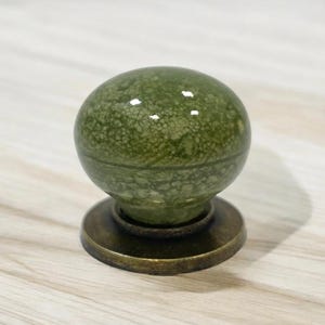 Peut inclure: Un bouton de meuble rond doté d'une surface en céramique vert olive brillant et marbré avec une texture craquelée. Le bouton est monté sur une base métallique circulaire de couleur bronze foncé avec une finition lisse et patinée.
