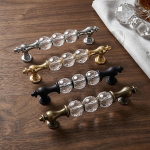 May include: Four cabinet pulls arranged in a row, each featuring three clear, faceted, spherical glass beads centered between two metal end posts. The metal finishes include polished chrome, bright gold, matte black, and an antique brass tone.