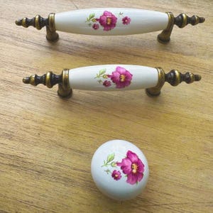 May include: Three decorative cabinet hardware pieces. Two are long, white porcelain handles with brass-colored ends and a floral design in pink and green. The third is a white porcelain knob with the same floral design.