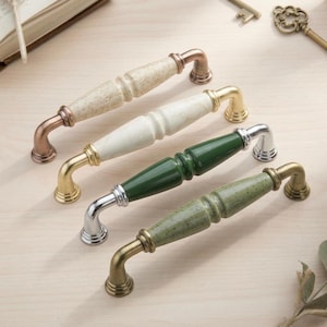 May include: Four cabinet pulls with ceramic handles in various colours: speckled beige, white, dark green, and speckled green. Each pull has a metal base in copper, gold, silver, and gold tones. The pulls are arranged on a light wood surface.