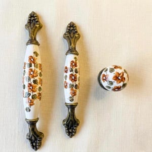 以下が含まれることがあります： ヴィンテージスタイルのキャビネット金具3点セット。白とブロンズの花柄のハンドル2つと、お揃いの丸いノブが含まれています。ハンドルは、ベースにブロンズのブドウの房のデザインと装飾的なトップが付いています。