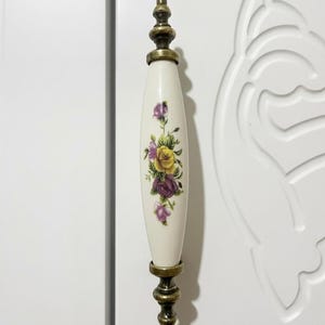 May include: A decorative door handle with a cream-colored ceramic center featuring a floral design in yellow, purple, and pink. The handle has brass-colored metal accents and is set against a white door.