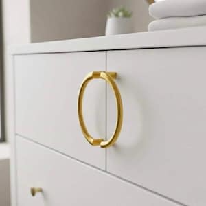May include: Close-up of a white dresser with gold-colored ring drawer pulls. The ring pulls are oval-shaped and attached to the white drawers. A smaller gold-colored knob is visible on the bottom drawer.