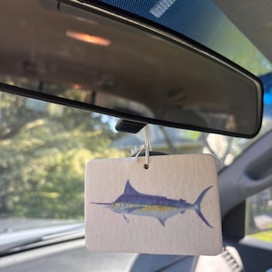 May include: A rectangular, off-white car air freshener hanging from a rearview mirror. The air freshener features a detailed illustration of a marlin fish with a blue back, a yellow stripe along its side, and a long, pointed bill extending forward.