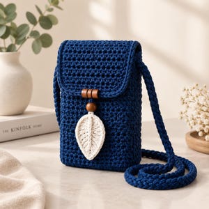 May include: A dark blue crochet crossbody bag featuring a rectangular shape with a flap closure. The bag is adorned with a central wooden bead and a cream-coloured crocheted leaf embellishment. It includes a thick, braided dark blue shoulder strap.
