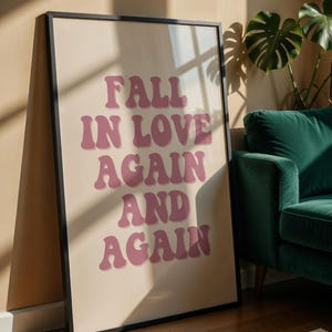 Könnte beinhalten: Ein gerahmter Kunstdruck mit dem Schriftzug "FALL IN LOVE AGAIN AND AGAIN" in rosa Retro-Lettern. Das Kunstwerk ist vor einem neutralen Hintergrund zu sehen und lehnt an einer Wand. Im Hintergrund sind ein grünes Sofa und eine Pflanze zu sehen.