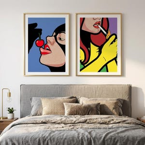 Pode incluir: Duas impressões de arte pop emolduradas com ilustrações estilizadas de mulheres. A impressão da esquerda mostra uma mulher com uma máscara preta segurando uma cereja vermelha perto dos lábios. A da direita mostra uma mulher ruiva acendendo um cigarro.