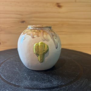 May include: A small, round ceramic vase with a light beige body and a green cactus design. The top rim has a brown and green glaze with drips. The vase is a decorative item.