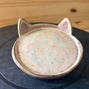 May include: A round ceramic trinket dish with two small pointed ears at the top. The dish features a speckled, mottled glaze in shades of pale pink, cream, and light blue, with a dark brown, textured clay rim visible around the edge.