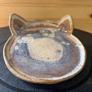 May include: A small, round ceramic dish shaped like a cat head with two pointed ears. The surface features a mottled glaze with blended tones of beige, brown, and deep blue, creating a textured, speckled appearance across the center and edges.