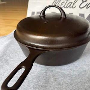 Può includere: Una padella in ghisa marrone scuro con coperchio. La padella ha un lungo manico con un foro e una base arrotondata. Il coperchio ha una maniglia ad anello. La padella è appoggiata su un panno grigio chiaro.