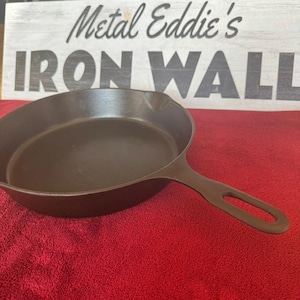 Puede incluir: Una sartén de hierro fundido marrón oscuro con un mango largo. La sartén está sobre una superficie roja. Encima de la sartén hay un letrero de madera que dice "Metal Eddie's IRON WALL".