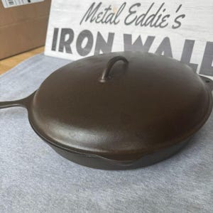 Könnte beinhalten: Eine dunkelbraune gusseiserne Bratpfanne mit passendem Deckel. Der Deckel hat einen gebogenen Griff. Die Bratpfanne steht auf einer grauen Oberfläche. Der Text "Metal Eddie's IRON WALL" ist im Hintergrund zu sehen.