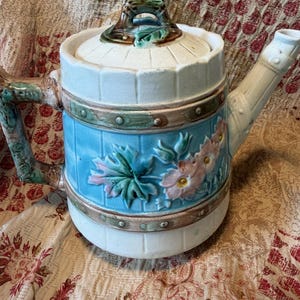 May include: A ceramic teapot shaped like a barrel with a light blue center band featuring embossed pink flowers and green leaves. The teapot has a white lid with a green handle, a brown handle on the side, and a white spout with a textured finish.