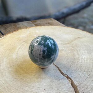 Op de afbeelding: Een gepolijste edelsteen bol, voornamelijk groen met witte en grijze insluitsels, rust op een natuurlijke houtsnipper. Het oppervlak van de bol reflecteert licht. De houtsnipper toont zichtbare groeiringen en een duidelijke scheur.