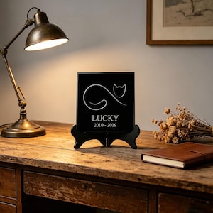 Puede incluir: Una placa conmemorativa cuadrada de color negro con un dibujo lineal blanco de un gato y el texto LUCKY 2010-2019. La placa se sostiene sobre un soporte de plástico negro, mostrando un diseño limpio con letras blancas claras y un gráfico.