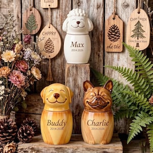 Puede incluir: Tres urnas funerarias de cerámica para mascotas con forma de perros sonrientes y una oveja, con nombres y fechas. Las urnas son amarillas, marrones y blancas. En el fondo cuelgan etiquetas de madera con diseños de temática natural.