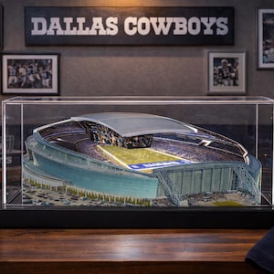 May include: A detailed 3D model of the Dallas Cowboys stadium enclosed in a clear rectangular acrylic display case with a black base. The miniature stadium features a retractable roof, a green football field, and surrounding parking lots.