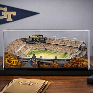 May include: A detailed diorama of a football stadium, encased in a clear display case. The stadium features a green field, seating, and a scoreboard. A pennant, mug, helmet, and notebook with the letters "GT" are also visible.