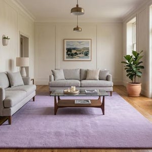 Könnte beinhalten: Ein geräumiges Wohnzimmer mit hellgrauen Sofas, einem Couchtisch aus Holz und einem lila Teppich. Zwei Topfpflanzen flankieren eine Glastür. Kunstwerke hängen an den Wänden, und Lampen stehen auf einem Beistelltisch.