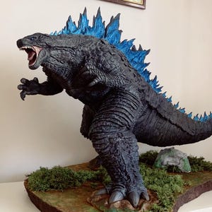 Può includere: Scultura dettagliata di Godzilla, un mostro immaginario, in posa dinamica con la bocca aperta. La figura è principalmente grigio scuro con pinne dorsali blu. La base presenta muschio verde e una piccola roccia, con il testo "Legendary Godzilla" sopra.