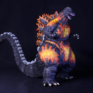 May include: A Godzilla action figure featuring a textured black body with vibrant orange and yellow glowing patterns across the chest, torso, and dorsal fins. The creature stands in a bipedal pose with a long, curved tail and sharp claws.