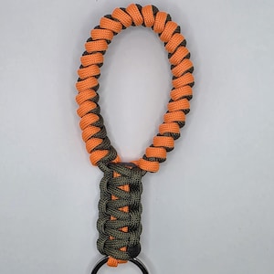 May include: A handmade keychain with a woven design. The keychain features orange and black paracord in a looped design, connected to a section of olive green and black paracord, and a black metal keyring.