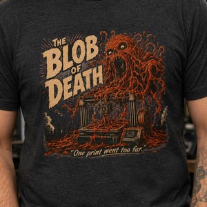 May include: A charcoal gray short-sleeve t-shirt featuring a graphic print of a monstrous orange blob consuming a 3D printer. The text reads THE BLOB OF DEATH and One print went too far. The design uses a distressed, vintage-inspired aesthetic.
