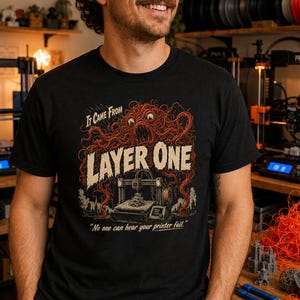 May include: Black t-shirt with the text "It Came From Layer One" and a graphic of a 3D printer and a monster. The shirt also has the text "No one can hear your printer fail." The person is smiling and standing in front of 3D printers and spools of filament.