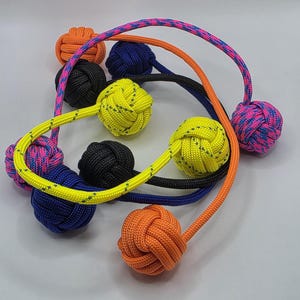 May include: A collection of colorful monkey fist knots with attached cords. The knots are in various colors, including orange, black, yellow, blue, and pink. The cords are also in matching or contrasting colors, creating a visually appealing display.