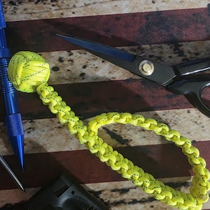 May include: A bright neon yellow paracord lanyard featuring a monkey fist knot at one end and a braided cord loop. The lanyard is positioned alongside a blue metal tactical pen, a pair of black scissors, and a black and orange butane torch lighter.