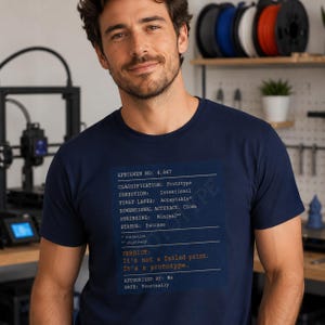 Op de afbeelding: Marineblauw t-shirt met een bedrukt label met de tekst "SPECIMEN NO: 4,847" en andere technische details. De tekst "It's not a failed print. It's a prototype" staat ook op het shirt. Het t-shirt wordt door een persoon gedragen.