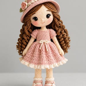 May include: A handmade crochet doll with brown curly hair, wearing a pink dress, hat, and shoes. The doll has a sweet face with rosy cheeks and large eyes. The dress has a white lace trim and a bow at the waist. The hat is decorated with a flower.
