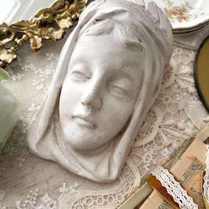 May include: A white plaster sculpture of a serene face with closed eyes, possibly a religious figure. The sculpture is set on a lace-covered surface, with vintage books and decorative plates in the background. The sculpture is a light gray color.