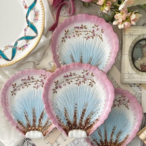 May include: A set of four ceramic dishes shaped like scallop shells, featuring a gradient of pale blue at the center transitioning to a scalloped pink rim. Each dish is decorated with hand-painted floral sprigs and small raised gold-tone accents.