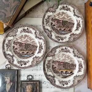 May include: Three round ceramic plates with scalloped edges, featuring a brown transferware landscape print of a stone bridge, trees, and buildings. The border has a floral pattern. Two small framed portraits and a wooden tool are also present.