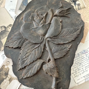 May include: A dark brown, weathered metal plaque featuring a high-relief sculpture of a single rose in full bloom with a smaller rosebud, detailed leaves, and a thorny stem. The surface has a textured, aged patina with visible metallic highlights.