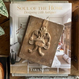 May include: A decorative wooden panel with a carved fleur-de-lis design rests on a book titled "Soul of the Home: Designing with Antiques" by Tara Shaw. The panel and book are on a wooden surface.