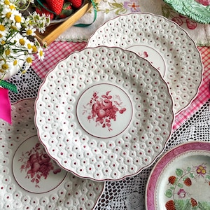 May include: A collection of white ceramic plates with a red floral design in the center and around the rim. The plates have a textured, dotted pattern. Strawberries and flowers are arranged nearby, suggesting a theme of spring or summer dining.