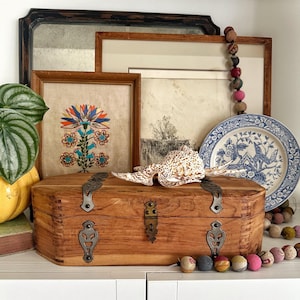 May include: A wooden chest with metal hardware is the focal point of this still life. A seashell rests on top. Behind the chest are framed pictures, a blue and white plate, and a decorative garland. The chest is approximately 12 inches long.