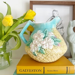 May include: A ceramic pitcher with a light blue top and handle, and a basket-weave design on the body. White and pink floral accents adorn the front. The pitcher sits atop two books, one yellow and one turquoise, with the titles "GALVESTON" and "THE BEAST".