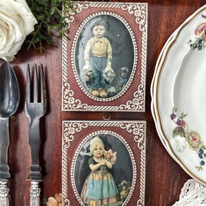May include: Two framed vintage illustrations of children. The top frame features a boy in a blue and yellow outfit holding a bouquet of flowers. The bottom frame features a girl in a blue and white dress holding a bouquet of flowers. Both frames have ornate silver trim and a red background.
