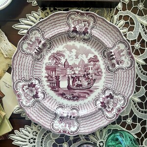 May include: A decorative antique plate with a maroon and white design. The plate features a central scene of people in a boat, surrounded by floral and architectural motifs. The rim has a wavy pattern and floral accents. The plate is approximately 10 inches in diameter.